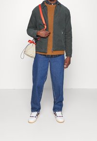 Groene cordjacket over een bruine gebreide trui, blauwe denim broek en witte sneakers met rode en blauwe accenten. Beige tas met een oranje schouderband.