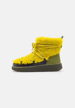 Botină cu blană galbenă, cu accente din piele verde, având o talpă masivă și șireturi. Designul include un vârf rotund și o suprafață texturată.