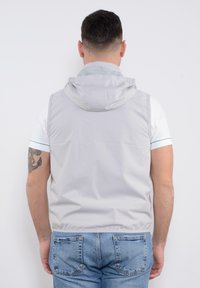 Gilet senza maniche grigio chiaro con cappuccio, caratterizzato da una texture liscia e un orlo elastico. Indossato sopra una camicia bianca con accenti blu e jeans.