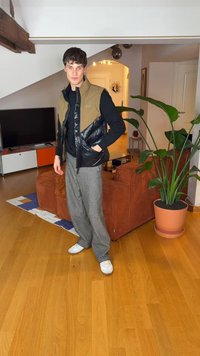 Juoda ir ruda pūsta liemenė dėvima ant juodo megztinio, pilkos plataus kirpimo kelnės ir balti sportbačiai. Vidaus aplinka su augalu ir baldais.