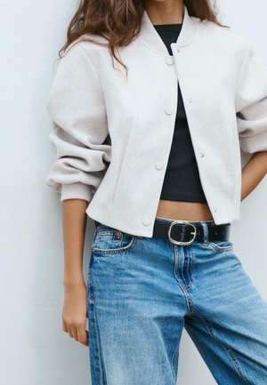 Femme portant une veste courte gris clair boutonnée, un haut court noir, un jean bleu taille haute et une ceinture noire avec une boucle argentée.