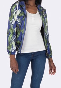 Letervægt blå jakke med tropisk bladmønster, lynlåslukning, elastiske ærmer og hætte, båret over en hvid bluse og blå jeans.