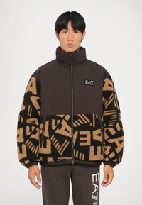 Ruda zip-up striukė su aukštu apykakle, kontrastuojančia pūkuota medžiaga su ryškiais raštais juodos ir smėlio spalvos, bei EA7 logotipu ant krūtinės.