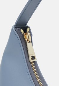 Vaaleansininen nahkabäg, jonka kulmassa on kultainen vetoketju ja Furla-logo; sileä pinta ja kontrastoiva harmaa kangasvuori.