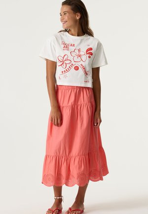 Camiseta blanca corta con gráficos florales rojos combinada con una falda larga coral que presenta un dobladillo de encaje con ojales y un diseño de capas.