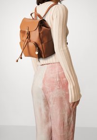 Zaino in pelle marrone con una patta pieghevole, chiusura con coulisse, tasca frontale con zip e un charm attaccato. Maglione beige a coste e pantaloni rosa chiaro.