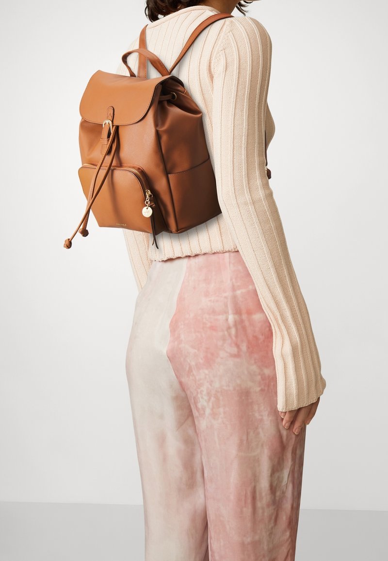 Zaino in pelle marrone con una patta pieghevole, chiusura con coulisse, tasca frontale con zip e un charm attaccato. Maglione beige a coste e pantaloni rosa chiaro.