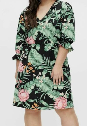Vrouw draagt een zwarte jurk met groene tropische bladeren en roze bloemmotieven, met driekwart gerimpelde mouwen en een V-hals.