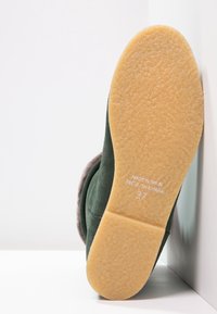Pantoufles en daim vert avec une doublure grise douce, semelle en caoutchouc texturé de couleur tan. Présente le texte en relief "FABRIQUÉ EN ESPAGNE" et la taille "27" au dos.