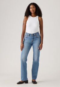 Jeans bleus taille haute avec une coupe décontractée, associés à un débardeur blanc à côtes. Des ballerines noires avec un petit talon accentuent la tenue.
