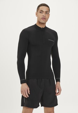 Schwarzes langärmeliges Sportshirt mit einem Halbreißverschluss-Design, strukturiertem Stoff und dezentem Branding. Kombiniert mit schwarzen Shorts.