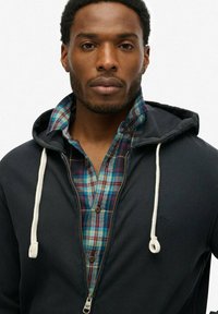 Sudadera negra con cremallera y cordones sobre una camisa a cuadros azul y verde con cierres de botones. Tela suave con capas texturizadas visibles.