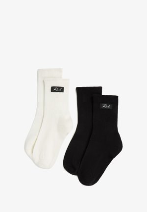 Deux paires de chaussettes côtelées : une en blanc cassé avec une étiquette de logo noire, et une en noir uni, toutes deux présentant un design simple et fonctionnel.