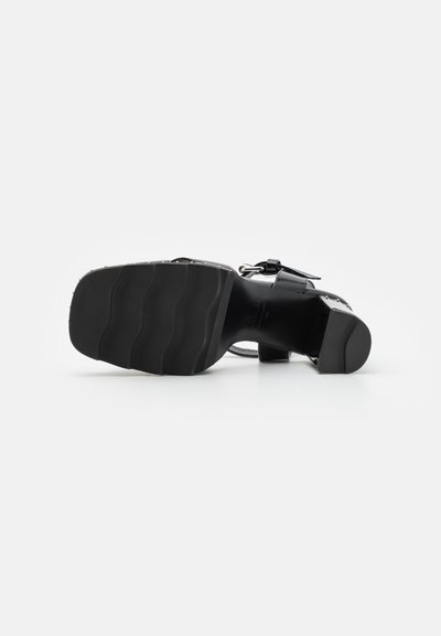 Zapato de tacón alto negro con suela de goma texturizada y tacón grueso, con una correa de hebilla y acentos metálicos en contraste.