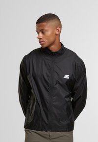 Giovane uomo che indossa una giacca nera con zip e un piccolo logo bianco sul petto sinistro, che guarda a sinistra su uno sfondo grigio uniforme.