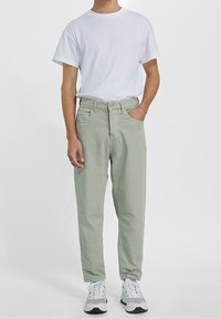 Pantalones de algodón verde claro con un corte relajado, bolsillos frontales y cierre de botón, combinados con una camiseta blanca básica y zapatillas grises.