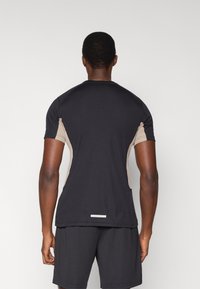 Μαύρο αθλητικό μπλουζάκι με μπεζ πλαϊνές λωρίδες, κοντά μανίκια και ίσιο τελείωμα. Διαθέτει ανακλαστικό στοιχείο κοντά στο κάτω μέρος.