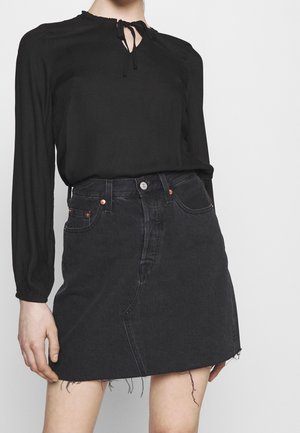 Femme portant un chemisier noir à manches longues avec un col à nouer et une mini-jupe en jean noire taille haute avec un ourlet effiloché.
