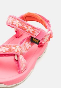 Sandalia Teva rosa con correas ajustables de patrón floral y suela acolchada, con una etiqueta de logo Teva en negro y amarillo.