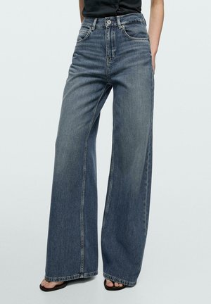 Jean denim bleu à jambes larges avec taille haute, bouton et fermeture éclair à l'avant, porté avec un haut noir et des sandales noires à bout ouvert.
