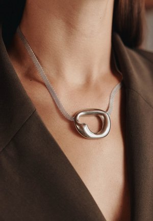 Zilveren metalen halsketting met een grote, sculpturale hanger, bevestigd aan een maas ketting, gedragen op een donkere stoffen achtergrond.