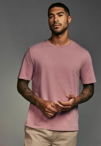 Tricou cu mânecă scurtă, guler rotund, din bumbac roz deschis. Prezintă o croială obișnuită și o textură netedă, afișat cu mâinile odihnindu-se în față.