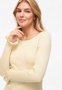 Jeune femme aux cheveux blonds portant un pull crème côtelé avec des poignets à volants, reposant le menton sur la main et souriant doucement sur un fond uni.