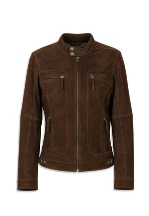 Homme portant une veste en daim marron, une chemise blanche, un pantalon noir large et des baskets, debout les mains dans les poches, regardant sur le côté.