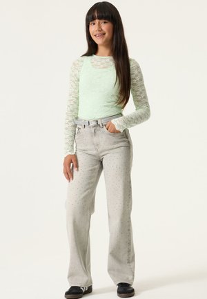 Tienermeisje staat met één hand in de zak, draagt een mintgroene kanten top met lange mouwen en lichtgrijze spijkerbroek met polkadots, gecombineerd met zwarte sneakers.