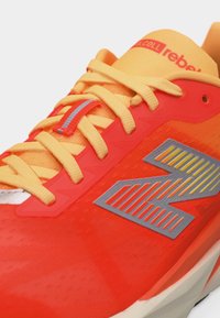 Φωτεινό πορτοκαλί και κίτρινο αθλητικό παπούτσι με κίτρινα κορδόνια, ανακλαστική λωρίδα στο βρόχο των κορδονιών και γκρι στυλιζαρισμένο λογότυπο "N" στο πλάι.