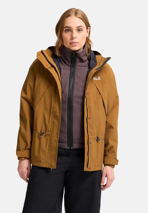 Vrouw draagt een bruine Jack Wolfskin-jas met capuchon over een donker vest met rits en zwarte broek, staand tegen een effen achtergrond.