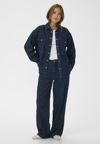 Donkerdenim jack met twee voorzakken, gecombineerd met loszittende donkerdenim broek. Voorzien van zilveren knopen en een schone, gladde textuur.