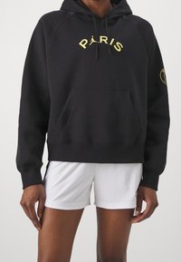 Sweat à capuche noir avec inscription jaune "PARIS", poche kangourou, poignets et ourlet côtelés. Short blanc complète la tenue. Style texturé et décontracté.