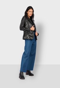 Černá kožená bunda s klopovým límcem, zapínáním na jeden knoflík a jemným leskem, spárovaná s širokými modrými džíny a robustními botami na podpatku.