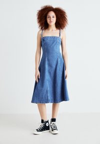 Denim A-linija haljina s podesivim trakama, svijetloplave boje, do koljena, s uskim tijelom i raširenom suknjom, uparenim s crnim tenisicama visoke pete.