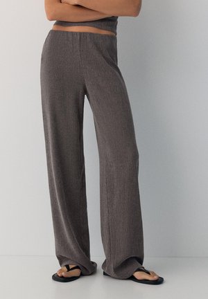 Person trägt lockere braune strukturierte Hose und schwarze Flip-Flops, steht mit verschränkten Armen vor einem schlichten hellen Hintergrund.