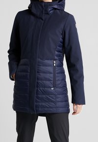 Marinblå isolerad kappa med huva, som har en kombination av släta och quiltade paneler, två fickor med dragkedja och en strömlinjeformad design.