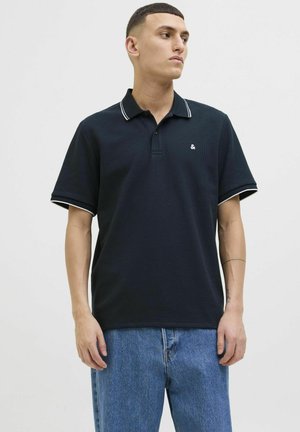 Dunkles Polohemd aus strukturiertem Stoff, mit weißem Besatz am Kragen und an den Ärmeln, ausgestattet mit einem kleinen Logo auf der linken Brust. Kombiniert mit blauen Jeans.