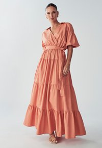 Long, tiered maxi dress in soft coral fabric. Features short sleeves, a wrap design, and a tied belt at the waist. Smooth texture.