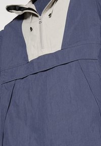 Navy and cream windbreaker with a half-zip collar, featuring a large front pocket and textured fabric. Accented with drawstring details.