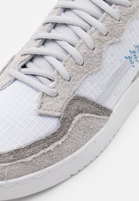 Baskets blanches avec des accents en suède gris texturé, dotées d'un design en tissu matelassé, de lacets gris et d'une semelle en caoutchouc blanche.