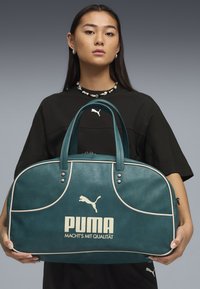 Ung kvinde holder tealfarvet Puma rejsetaske med cremefarvede accenter og logo, iført sort Puma-outfit og perlekæde mod grå baggrund.