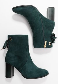 Stivaletti ankle in suede verde con tacco a blocco, cerniera laterale e dettaglio decorativo a fiocco. Tessuto liscio e design della punta arrotondata.
