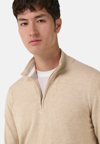 Pull beige à fermeture éclair avec une finition texturée, comportant un léger col et un accent rayé au niveau du cou. Porté par-dessus une chemise blanche.