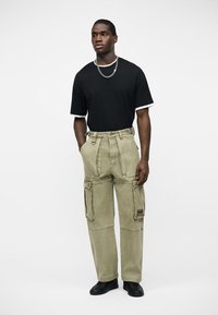 Άνδρας που στέκεται φορώντας μαύρο μπλουζάκι, αλυσίδα λαιμού από ασημί, μπεζ παντελόνι cargo με πολλές τσέπες και μαύρα παπούτσια μπροστά σε απλό φόντο.