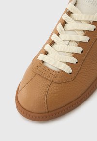 Καφέ δερμάτινο αθλητικό παπούτσι με υφή, κρεμ κορδόνια, στρογγυλεμένη μύτη και λαστιχένια σόλα με ζιγκ-ζαγκ μοτίβο κατά μήκος της άκρης.