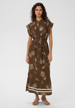 Nő áll, barna, bokáig érő ruhát visel, melyen krémszínű tengeri kagyló- és korallminta látható, megkötős derékrésszel és rövid, vállra simuló ujjakkal.