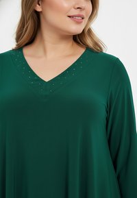 Groene langemouwen top met een V-halslijn en decoratieve studs langs de rand, gemaakt van zachte, lichte stof.