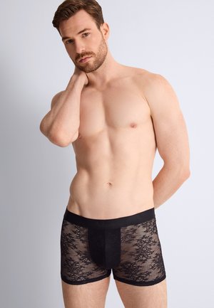 Homme torse nu avec une barbe portant un boxer en dentelle noire, debout devant un fond gris clair, une main touchant son cou.