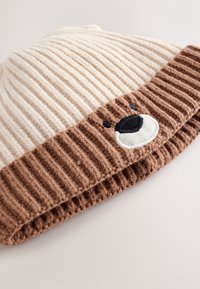 Kötött sapka bordázott mintával; krémszínű teteje és barna, texturált pereme hímzett medvearc és kör alakú orrdíszítéssel.